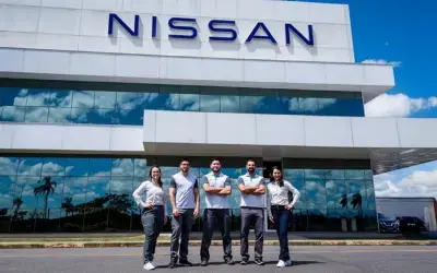Cinco funcionários da Nissan, usando uniformes com a marca, posam em frente a um prédio moderno de vidro. Eles estão em fila, com sorrisos e poses confiantes.