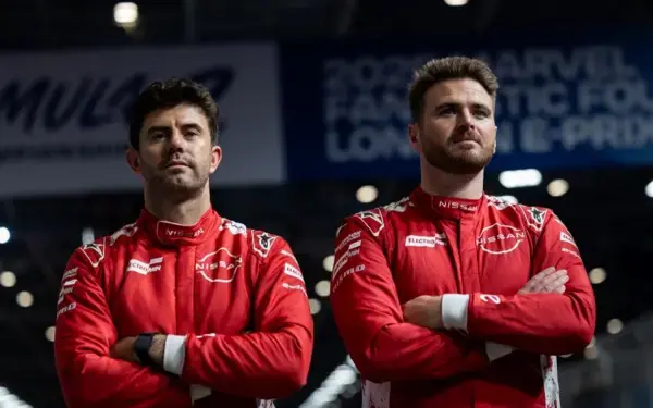 Dois pilotos da equipe de corrida da Nissan, usando macacões vermelhos com a marca, posam com os braços cruzados em um ambiente de pista de corrida.