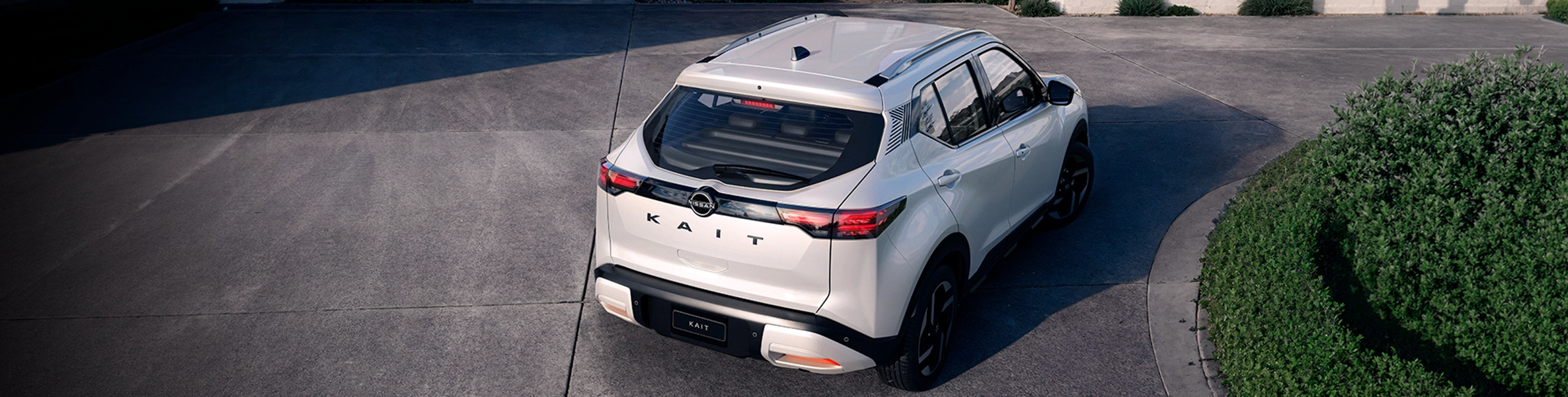 Vista superior traseira de um SUV Nissan branco a circular numa área pavimentada ao lado de um arbusto verde.