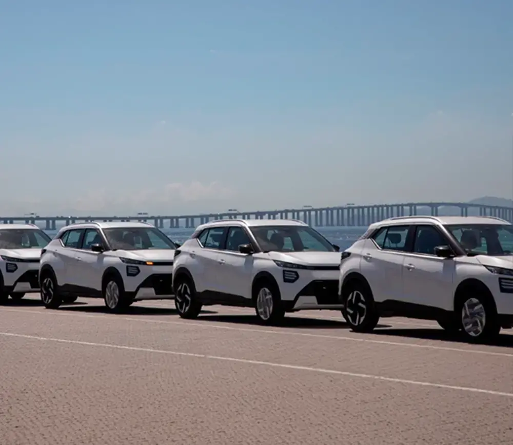 Fileira de carros Nissan Kait brancos alinhados em um estacionamento com uma ponte e o mar ao fundo.