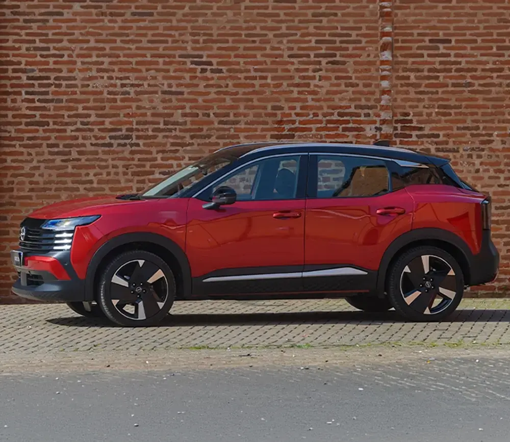 Visão lateral de perfil do SUV Nissan Kicks vermelho com teto preto estacionado em frente a uma parede de tijolos à vista.