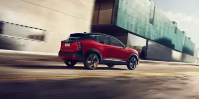 Nissan Kicks vermelho com teto preto em movimento por uma rua urbana, visto pelo ângulo traseiro direito com efeito de desfoque para indicar velocidade.