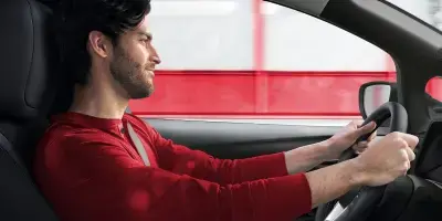 Homem de camisa vermelha segurando o volante de um carro em movimento, com um borrão de velocidade vermelho e branco visível fora da janela.