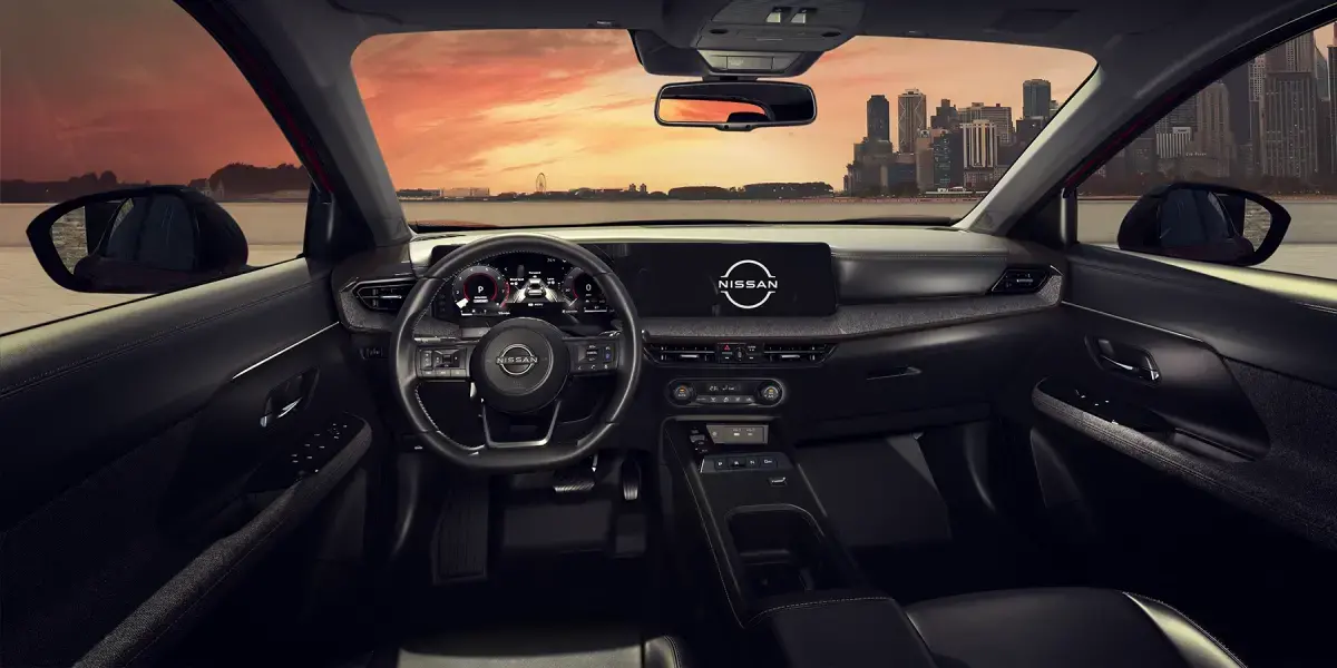 Interior do painel escuro de um carro Nissan Kicks, mostrando o volante e as telas digitais ligadas, com vista pelo para-brisa para uma cidade ao pôr do sol.