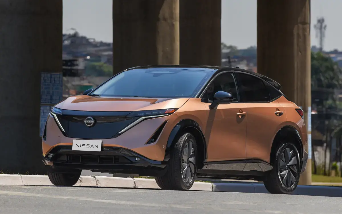 O Nissan Ariya elétrico na cor cobre com teto preto está parado em uma rua, com colunas de viaduto ao fundo. O design moderno e elegante do SUV elétrico destaca-se sob a luz do dia.