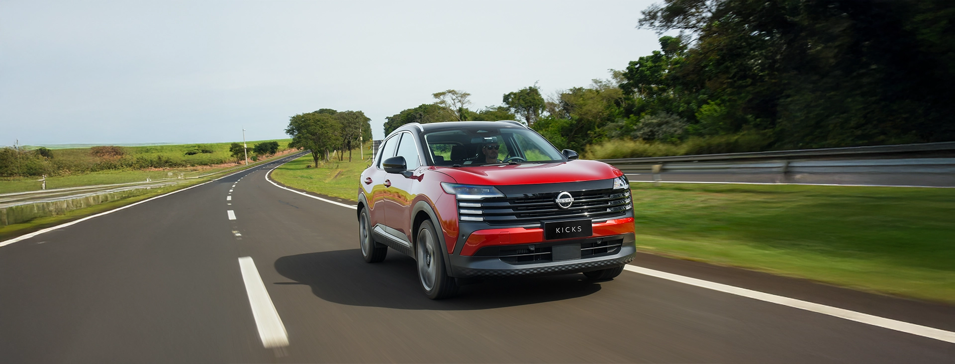 O SUV Nissan Kicks vermelho com teto preto dirige em uma estrada com curvas em meio a uma paisagem rural. O carro está em movimento, com o fundo borrado, destacando seu design.