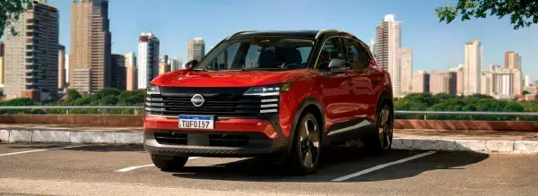 Nissan Kicks SUV vermelho estacionado em pátio, com a silhueta de arranha-céus da cidade em um dia ensolarado.