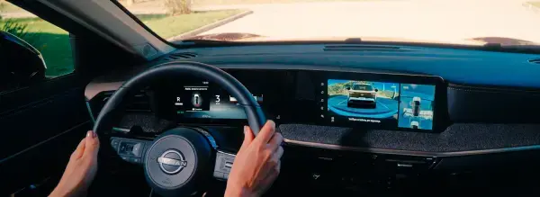 Painel do carro com volante e mãos do motorista. A tela de multimídia exibe a imagem da câmera de ré e a visão panorâmica de 360 graus do entorno do veículo.