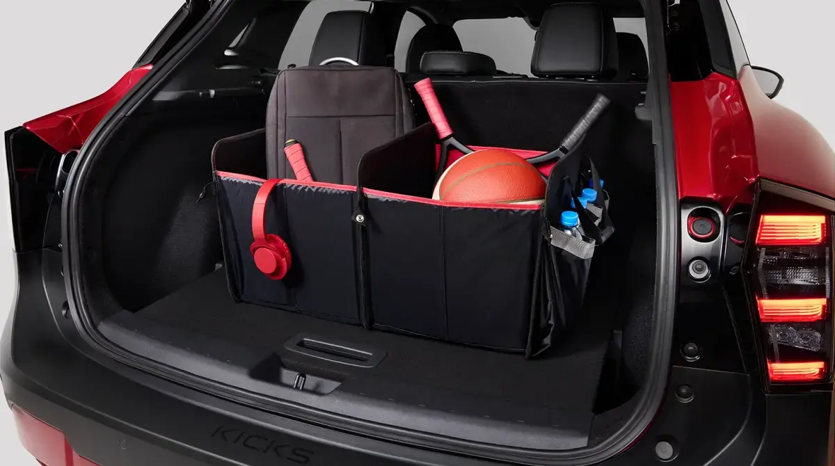 Uma mão segura um smartphone preto com o símbolo de Bluetooth branco na tela, posicionado à frente de um Nissan Kicks vermelho brilhante. O carro, com seu design frontal moderno e rodas com detalhes em preto e prata, está estacionado em um fundo claro.