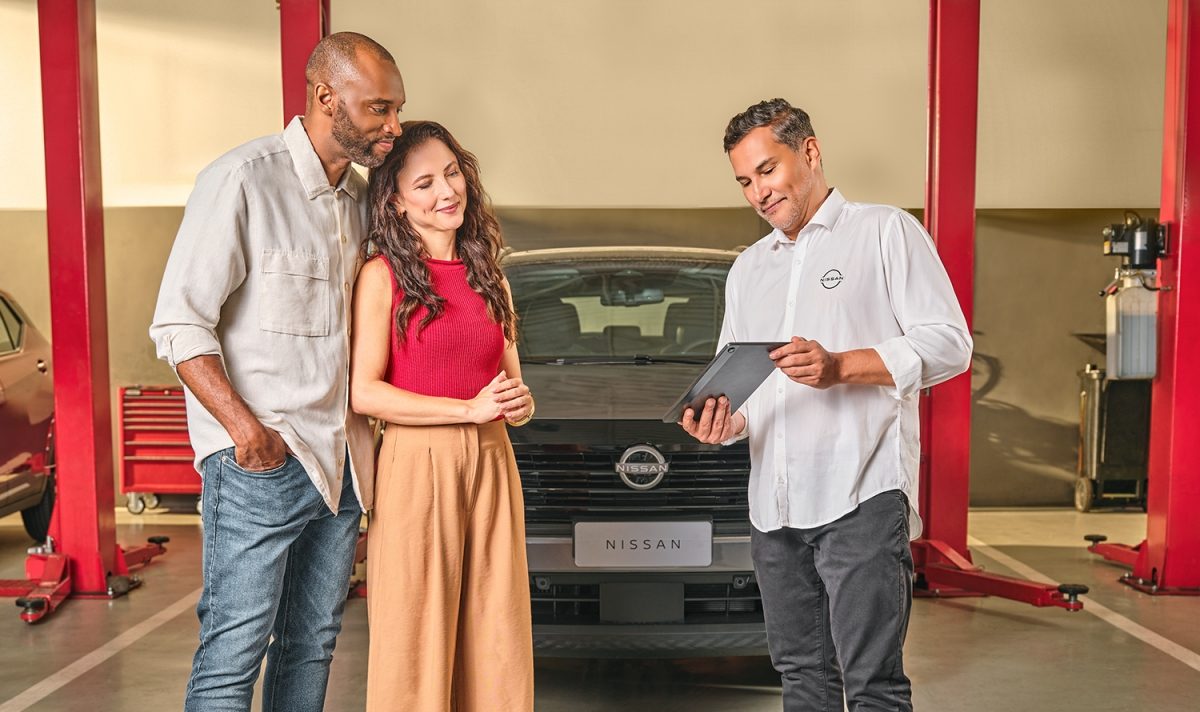 Consultor da Nissan mostrando informações em um tablet para um casal de clientes em uma oficina mecânica, com um carro da marca estacionado ao fundo.