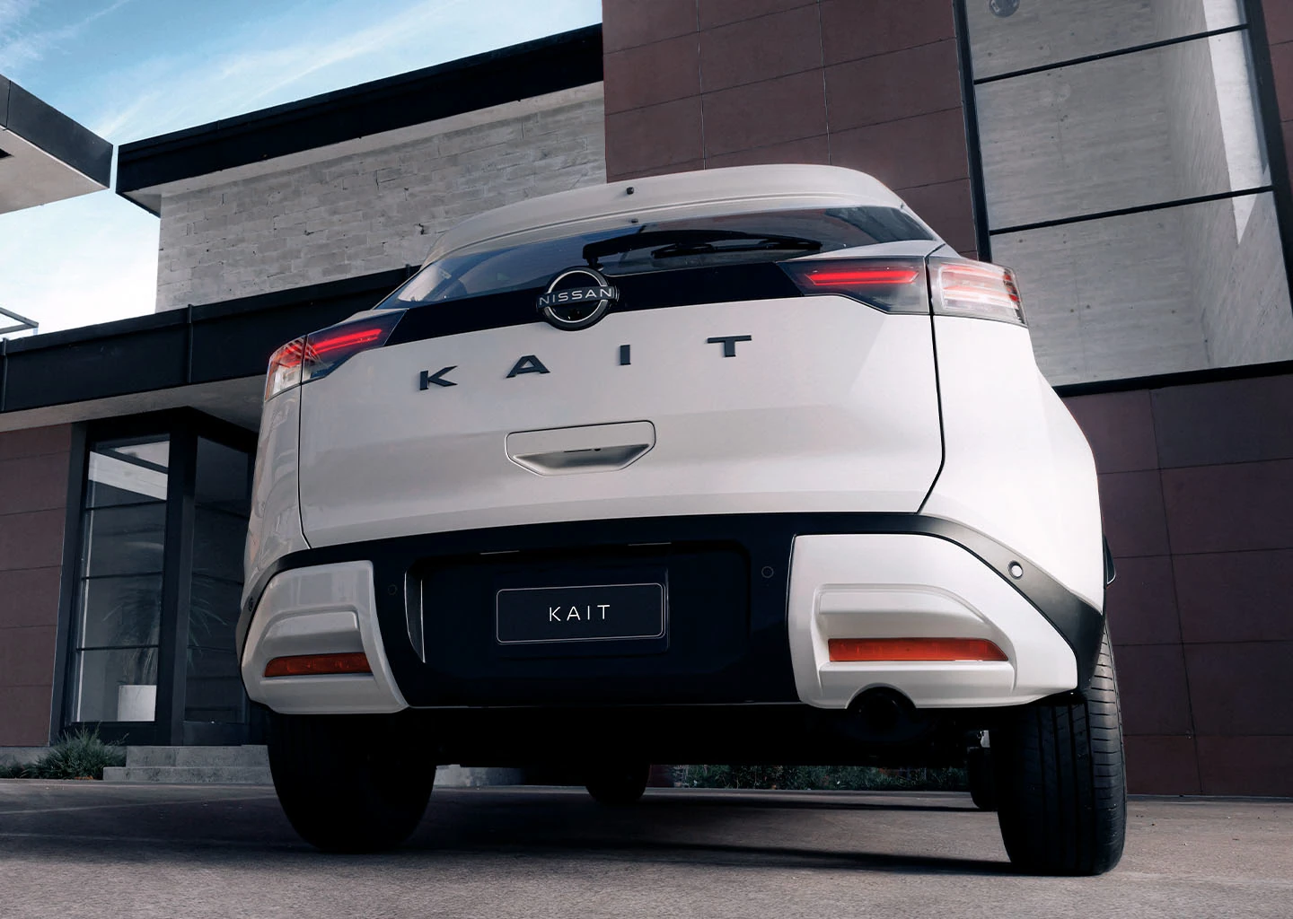 ista traseira de um Nissan KAIT branco estacionado em frente a uma residência de arquitetura moderna. As lanternas traseiras horizontais e o emblema "KAIT" são visíveis.