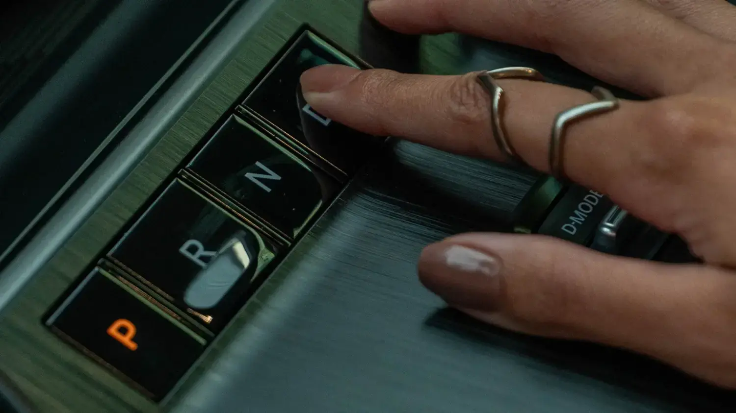 Close-up de uma mão com um anel tocando os botões do seletor de marchas eletrônico (E-shifter) de um Nissan Kicks, que inclui as opções "P", "R", "N" e "D". A superfície ao redor tem acabamento texturizado.