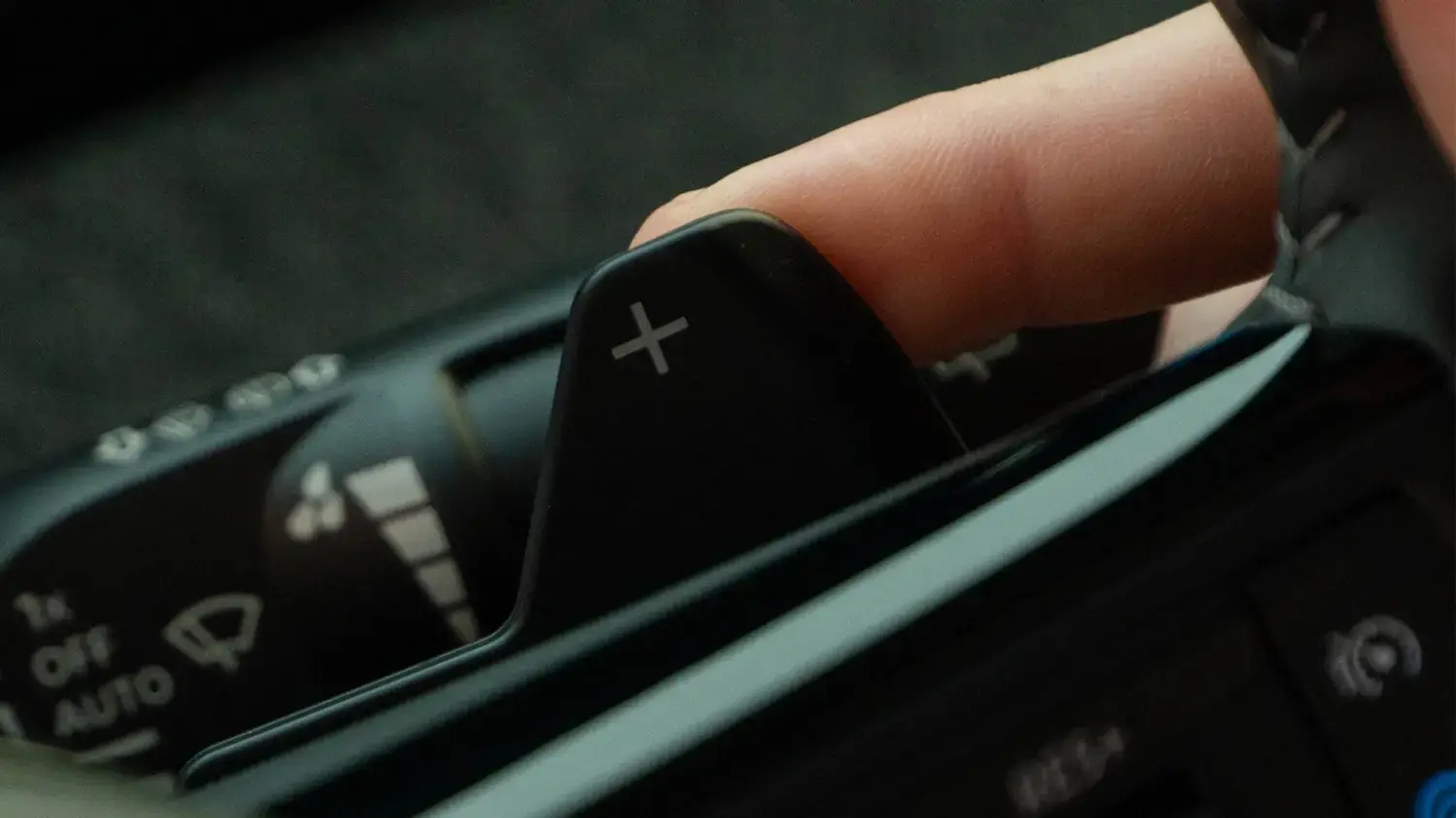 Close-up de um dedo tocando uma das alavancas de troca de marcha (paddle shifters) no volante de um Nissan Kicks, com o símbolo de "mais" (+) visível. Parte dos controles do limpador de para-brisa está ao fundo.