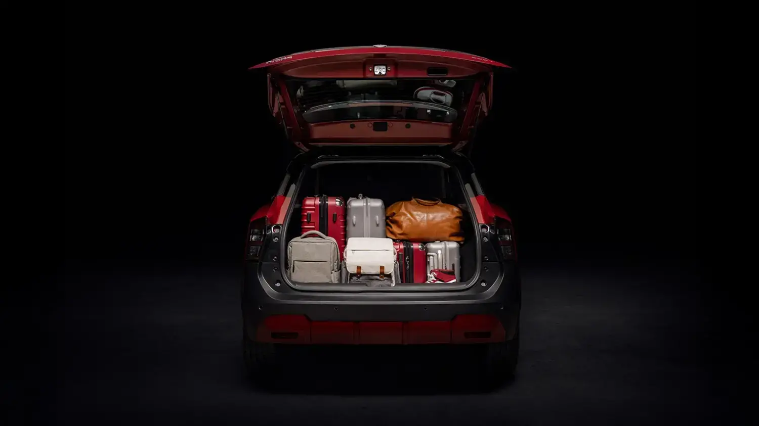 Vista traseira de um Nissan Kicks vermelho com a tampa do porta-malas aberta, revelando um amplo espaço de carga preenchido com várias malas de viagem de diferentes tamanhos e cores, sobre um fundo escuro.