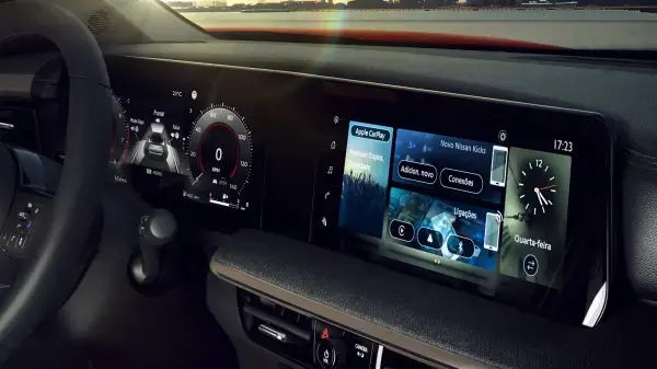 Painel digital duplo do Nissan Kicks, mostrando a tela do painel de instrumentos à esquerda com velocímetro e informações de direção, e a tela de multimídia à direita exibindo a interface do Apple CarPlay com opções de música, chamadas e um relógio digital. O interior do carro reflete a luz do pôr do sol.