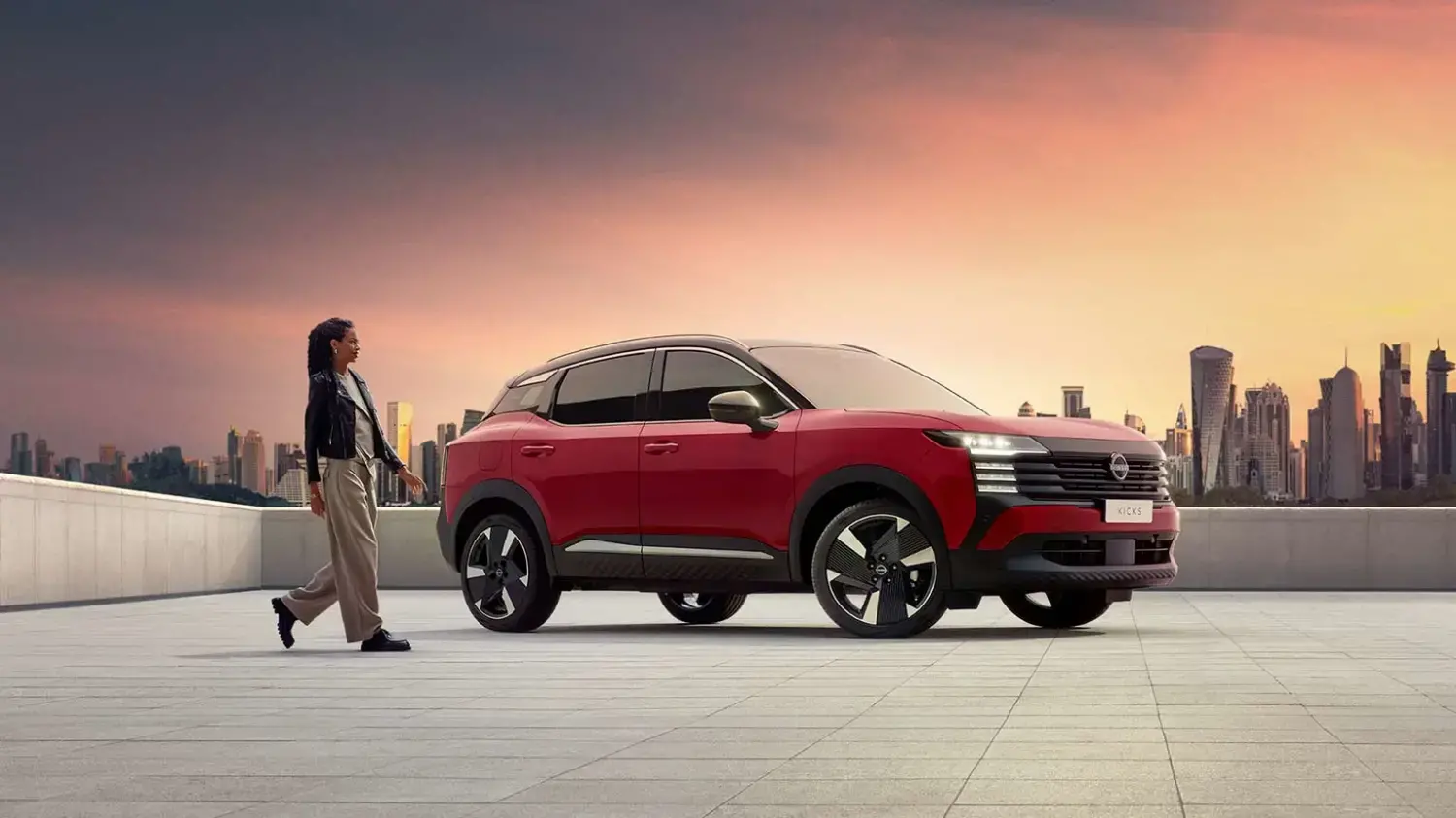 Uma pessoa com estilo casual caminha ao lado de um Nissan Kicks vermelho em um cenário urbano moderno. O SUV está estacionado em um terraço com uma silhueta de arranha-céus ao fundo e o céu em tons de pôr do sol, transmitindo um estilo de vida cosmopolita.