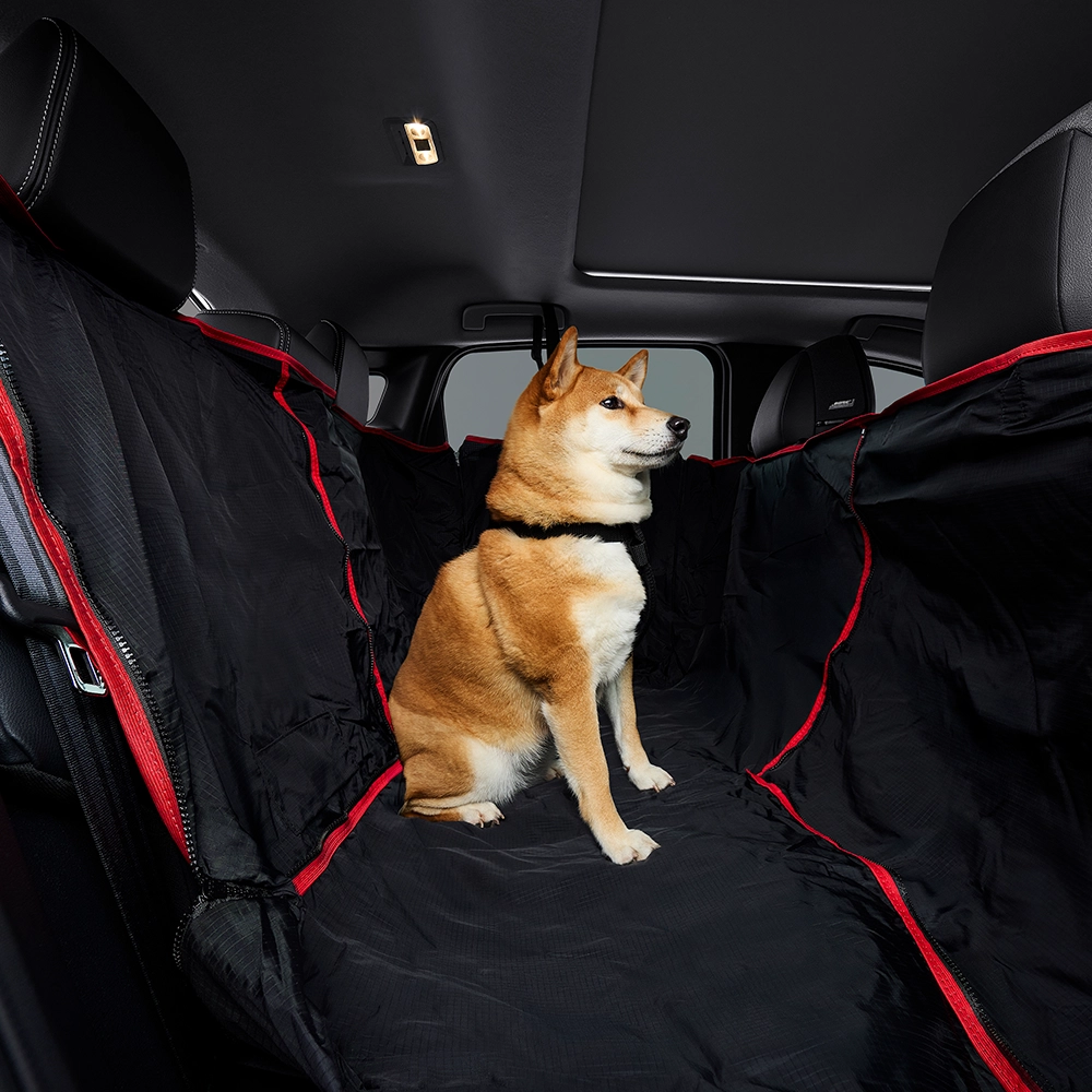 Um cachorro da raça Shiba Inu de pelagem marrom e branca está sentado no banco traseiro de um Nissan Kicks, que está equipado com uma capa protetora preta com detalhes em vermelho cobrindo os assentos. O teto solar do veículo é visível ao fundo.