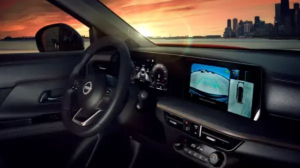 Interior de um Nissan Kicks com o painel digital aceso, mostrando a tela central de multimídia com uma visão de câmera 360 graus do entorno do carro e uma perspectiva do veículo visto de cima. O volante é visível no lado esquerdo e o pôr do sol se reflete no para-brisa.