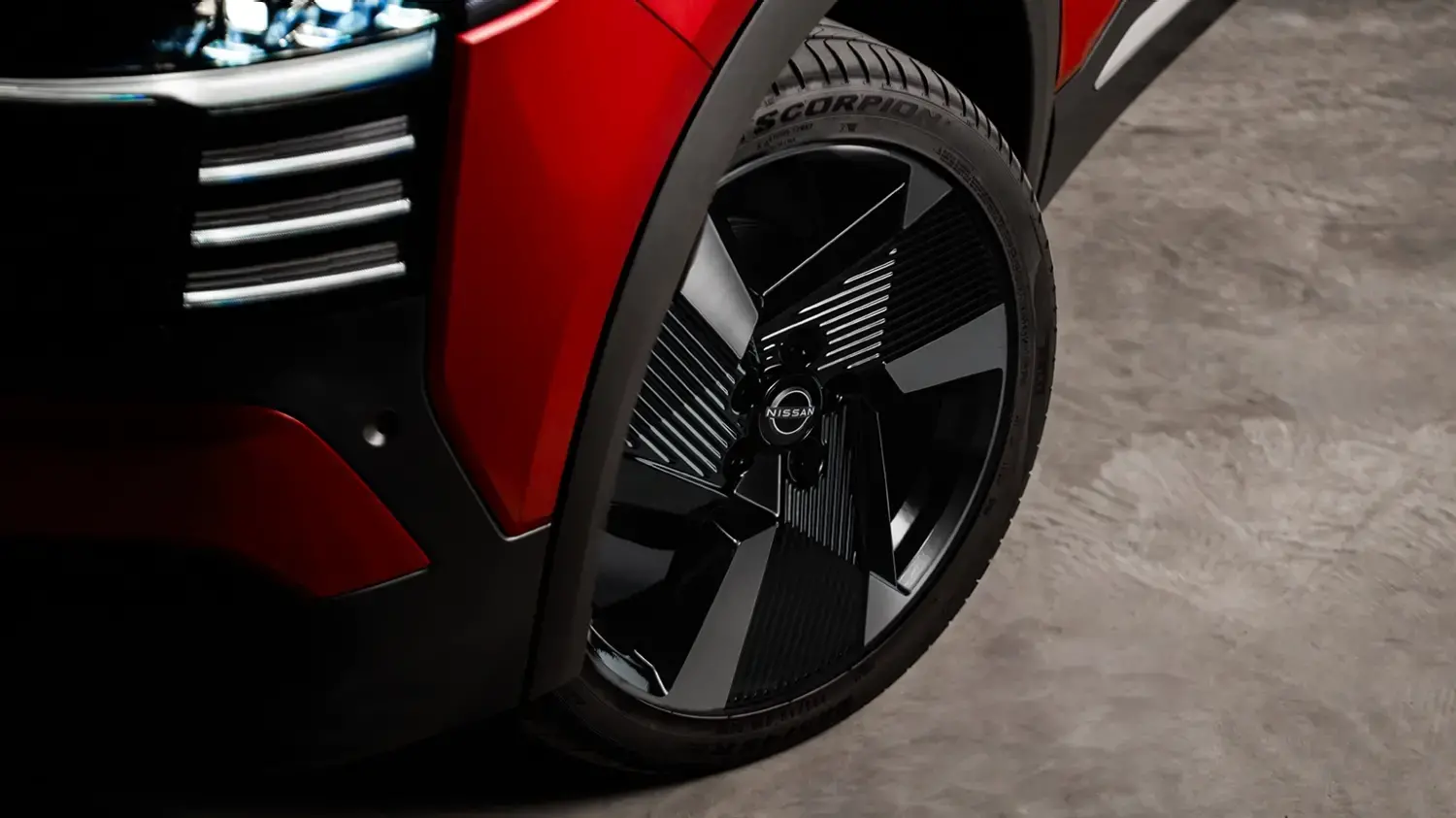 Close-up de uma roda de liga leve do Nissan Kicks vermelho, com design esportivo em preto e prata e o logotipo da Nissan ao centro. Pequenas partículas de luz brilhante ao redor da roda enfatizam o dinamismo do veículo.
