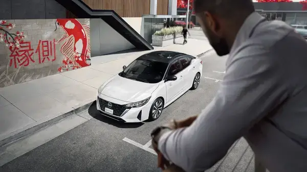 Chave de carro em cima de uma mesa de madeira, com um origami vermelho ao lado e uma pessoa ao fundo olhando para documentos.