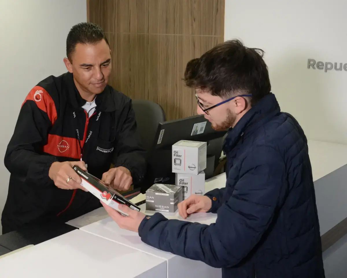 Vista lateral trasera 3/4 de Nissan URVAN en rampa de servicio y técnico de servicio ofreciendo diagnóstico de mantenimiento.