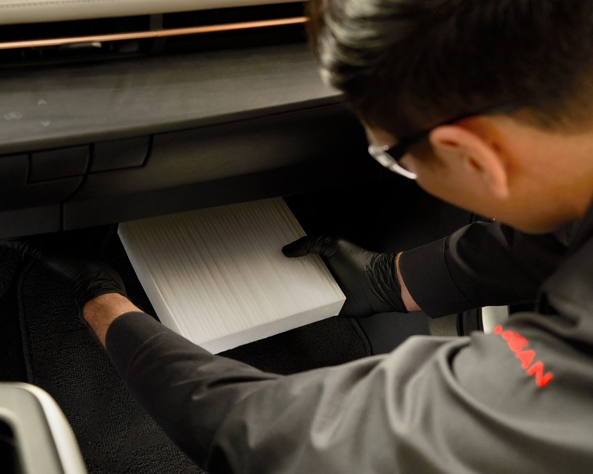 Vista lateral trasera 3/4 de Nissan URVAN en rampa de servicio y técnico de servicio ofreciendo diagnóstico de mantenimiento.